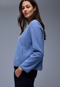 Vrouw met lang bruin haar, gekleed in een lichtblauw getextureerd jack met lange mouwen en een donkere broek, staand met één hand in de zak tegen een grijze achtergrond.