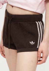Nahaufnahme einer Person, die schwarze, strukturierte Strickshorts mit weißen Streifen und Adidas-Logo an der Seite trägt, kombiniert mit einem hellrosa Oberteil.
