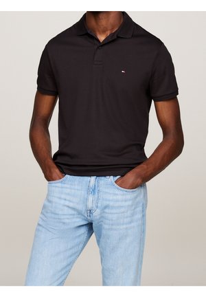 Mann trägt ein schwarzes kurzärmliges Polo-Shirt mit kleinem Logo und hellblauen Jeans, Hände in den Taschen, steht vor einem schlichten Hintergrund.