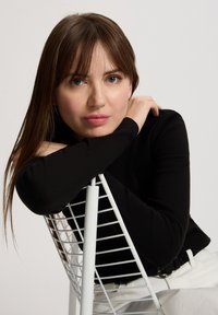 Czarna bluzka z długim rękawem noszona przez osobę opierającą się o biały metalowy fotel z siatkowym wzorem. Jasne tło, gładka tekstura, neutralne kolory.