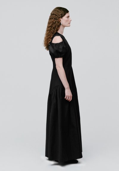 Frau mit langen lockigen Haaren, die ein schwarzes Maxikleid mit freiliegenden Schultern und Puffärmeln trägt, steht im Profil vor einem grauen Hintergrund.