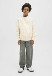 Krämfärgad sweatshirt med "ORIGINALS" präglat på framsidan, ihop med löst sittande grå jeans och svart-vita sneakers.