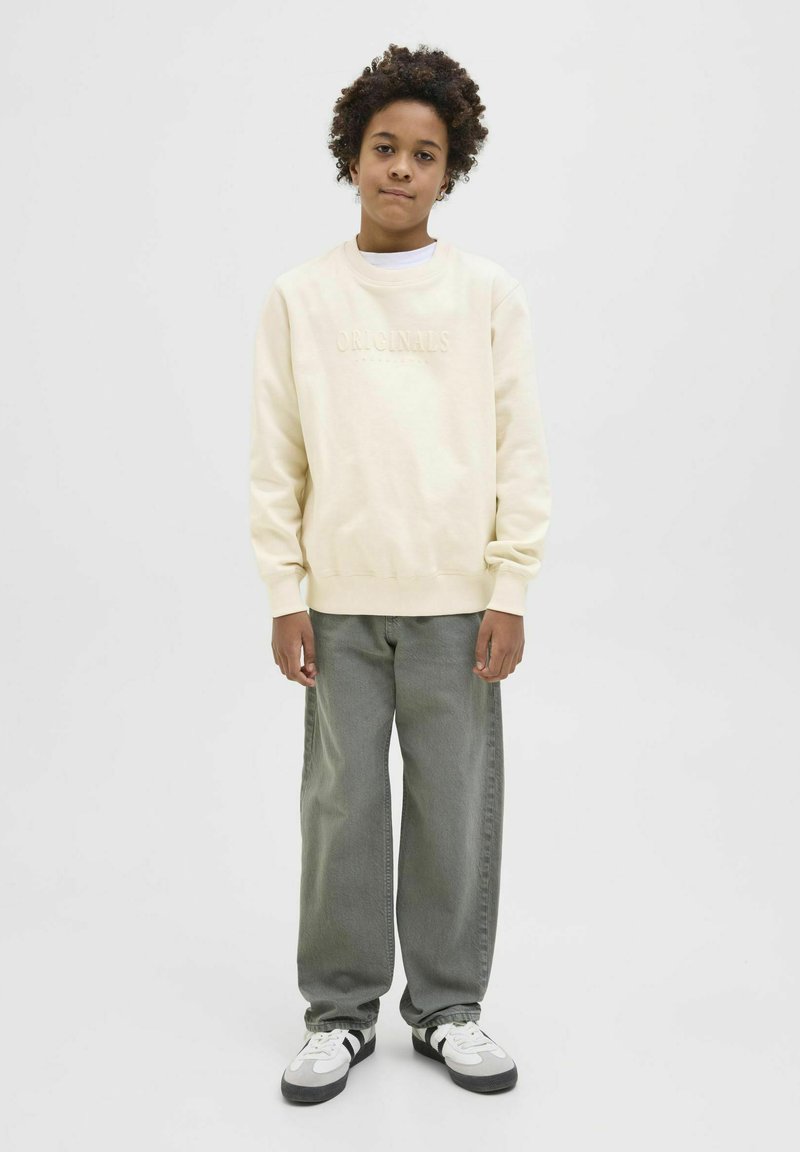 Krämfärgad sweatshirt med "ORIGINALS" präglat på framsidan, ihop med löst sittande grå jeans och svart-vita sneakers.