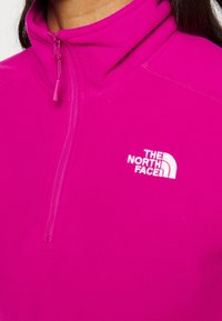 Personne portant un pull polaire rose vif avec une fermeture éclair quart de zip et le logo blanc The North Face sur la poitrine, fermeture éclair partiellement fermée près du cou.