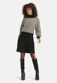 Gestreifter schwarz-cremefarbener Pullover mit langen Ärmeln, kombiniert mit einem schwarzen A-Linien-Rock mit Knopfdetaillierung an der Vorderseite und kniehohen schwarzen Stiefeln.