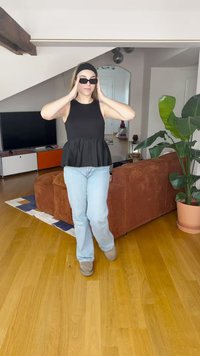 Svart peplum topp, lyseblå jeans, brune sko; iført svarte solbriller; innendørs setting med en plante og en brun sofa i bakgrunnen.