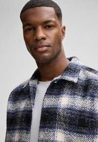 Kariertes Jackett in Blau, Schwarz und Weiß, aus strukturiertem Stoff gefertigt. Verfügt über einen Kragen und Knöpfe, getragen über einem grauen T-Shirt.