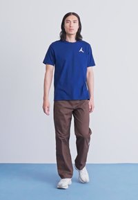 Bărbat cu păr lung și închis la culoare merge înainte, purtând un tricou Jordan de culoare bleu închis, pantaloni maro și adidași albi, pe un jos albastru și un fundal gri deschis.