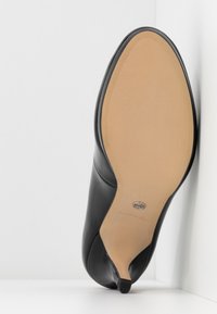 Chaussure à talon haut en cuir noir vue de côté, montrant une semelle beige lisse et un petit embout de talon sur une surface blanche.