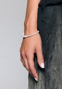 Bransoletka z koralików z okrągłymi białymi perłami o różnych rozmiarach oraz minimalistycznym złotym akcentem. Noszona na ręce na tle teksturowanej ciemnej tkaniny.