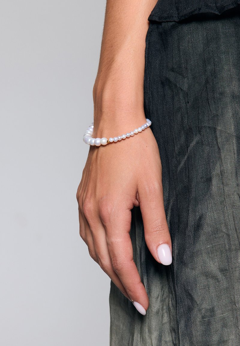 Bransoletka z koralików z okrągłymi białymi perłami o różnych rozmiarach oraz minimalistycznym złotym akcentem. Noszona na ręce na tle teksturowanej ciemnej tkaniny.