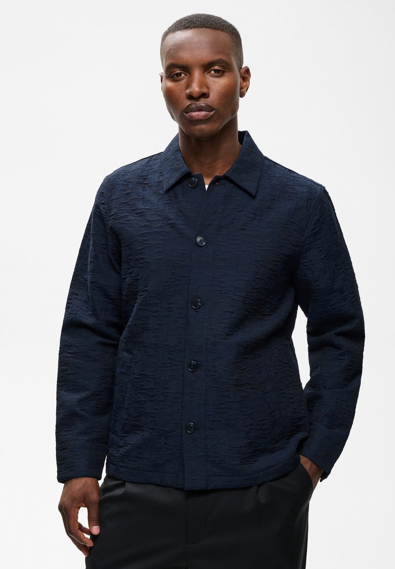 Homme aux cheveux courts portant une veste boutonnée texturée foncée avec col, debout une main dans la poche devant un fond clair uni.