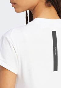 Bala t-krekls ar salocītām piedurknēm, aizmugurē ar vertikālu melnu svītru, kas ietver vārdu "SPORTA APĢĒRBS". Gluda auduma tekstūra.