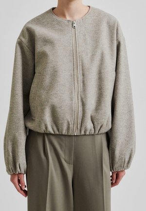 Osoba ubrana w jasnoszarą kurtkę na zamek z elastycznymi mankietami i dolnym brzegiem, zestawioną z oliwkowozielonymi spodniami z wysokim stanem.