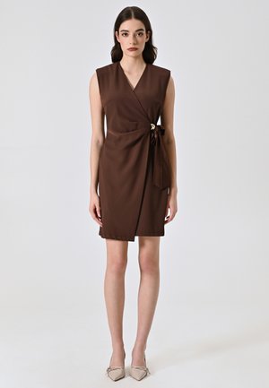 Robe brune à encolure en V profonde, sans manches, avec un détail de nœud sur le côté et une texture lisse. Portée avec des talons beiges.