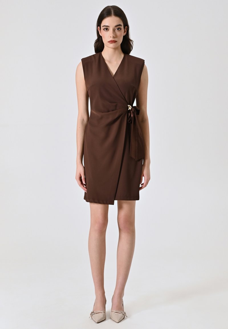 Braunes Wickelkleid mit tiefem V-Ausschnitt, ärmellosem Design, seitlichem Knoten-Detail und glatter Textur. Tragen mit beigen Absätzen.