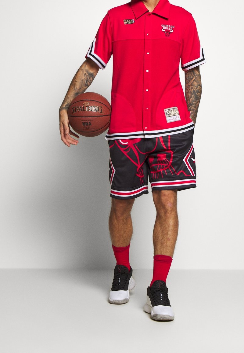 Chemise rouge à manches courtes avec boutons et accents noirs, ainsi qu'un logo de basketball, associée à un short noir avec un imprimé graphique rouge et des bandes blanches.