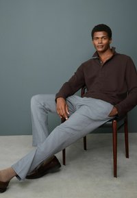 Dunkelbraunes Poloshirt, graue maßgeschneiderte Hose, sitzt auf einem Holzstuhl. Braune Schuhe und hellbraune Socken. Einfaches, sauberes Design, subtile Texturen.