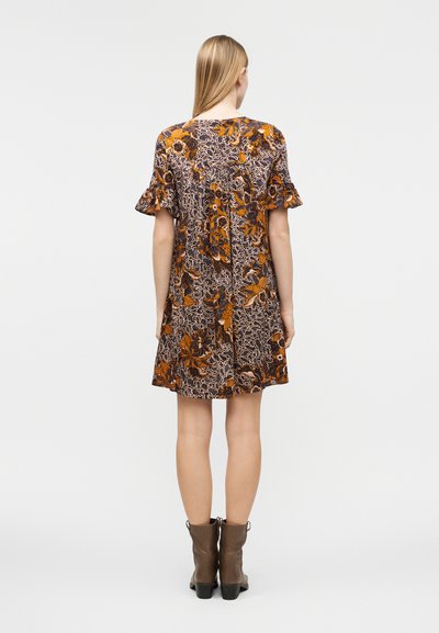 Γυναίκα με μακριά ξανθά μαλλιά, ντυμένη με κοντό φορεματάκι με λουλουδάτο σχέδιο και βολάν στα μανίκια, φορώντας καφέ μποτάκια μέχρι τον αστράγαλο, στέκεται γυρνώντας την πλάτη της.