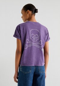 Lilla kortærmet t-shirt har et design med en nitteret dødningehoved og korslagte knogler på ryggen. Tekstureret bomuldsstof, afslappet pasform.