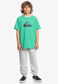 Camiseta de algodón verde con un logo de montaña negra, combinada con pantalones de chándal grises y zapatillas negras sin cordones. Atuendo simple y casual.