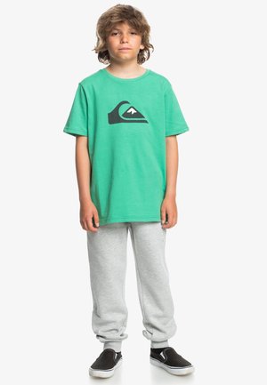 Camiseta de algodón verde con un logo de montaña negra, combinada con pantalones de chándal grises y zapatillas negras sin cordones. Atuendo simple y casual.