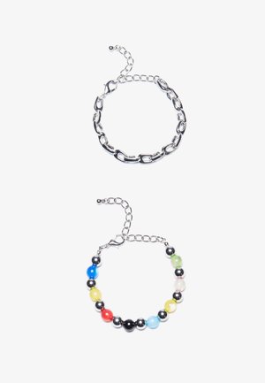 Ασημένια βραχιόλι αλυσίδας με επιμήκεις κρίκους από πάνω. Κάτω από αυτό, ένα πολύχρωμο βραχιόλι με χάντρες σε μαύρο, μπλε, πράσινο, κίτρινο, κόκκινο και διάφανο.
