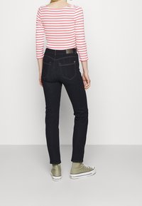 Jeans ajustados de mezclilla oscura con costuras naranjas, combinados con una camiseta de manga larga a rayas rojas y blancas. El conjunto incluye zapatillas en color verde oliva.