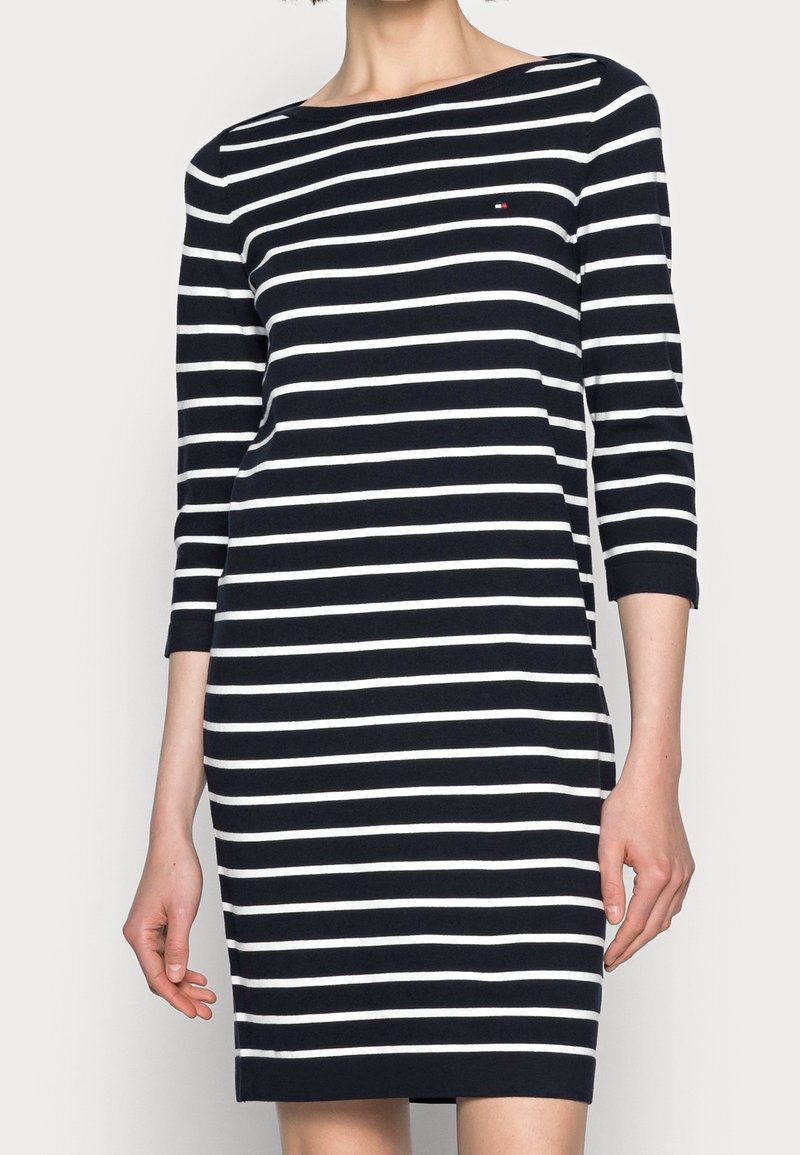 Robe rayée avec des rayures horizontales bleu marine et blanches, manches trois-quarts, encolure ronde, et un petit logo sur la poitrine. Tissu lisse.