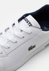 White leather sneaker with a textured surface, navy lining, white laces, and a green crocodile logo on the side. Lace-up closure.