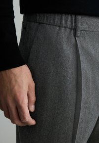 Graue Wollmischungshosen mit tailliertem Schnitt, ausgestattet mit Seitentaschen, flachen Vorderteilen und einer texturierten Oberfläche. Hand ruht auf dem Bund.