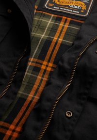 Sort jakke med en gylden lynlås; indvendigt har den et for i grønt, marineblåt og orange ternet mønster; mærkelogo synligt ved kraven.