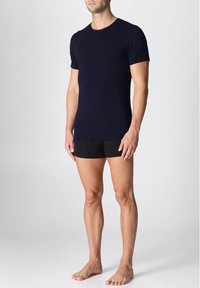 Uomo che indossa una t-shirt aderente a maniche corte blu scuro e boxer neri, in piedi a piedi nudi contro uno sfondo chiaro e semplice.