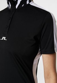 Sort polo-shirt med høj krave, med hvide detaljer på ærmerne og en kontrastfarvet mønstret hals. Knaplukning og logo-detalje.