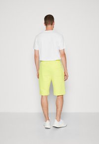 Gelbe Baumwollshorts mit einem elastischen Bund und einer Gesäßtasche, kombiniert mit einem schlichten weißen T-Shirt und weißen Sneakers.