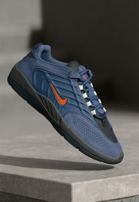 Modrá sportovní bota s texturovaným svrškem, černými akcenty, šedou podrážkou, výrazným oranžovým logem Nike a šněrováním.