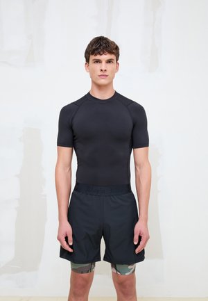 Joven con una camiseta negra ajustada de manga corta, pantalones cortos negros y mallas de compresión con estampado de camuflaje, de pie frente a un fondo claro y liso.