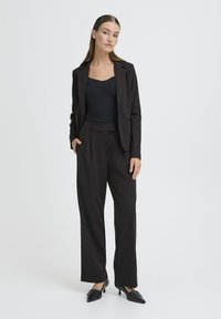 Donna in piedi con una mano in tasca, che indossa un blazer nero su misura, pantaloni larghi abbinati, top nero e décolleté neri a punta.