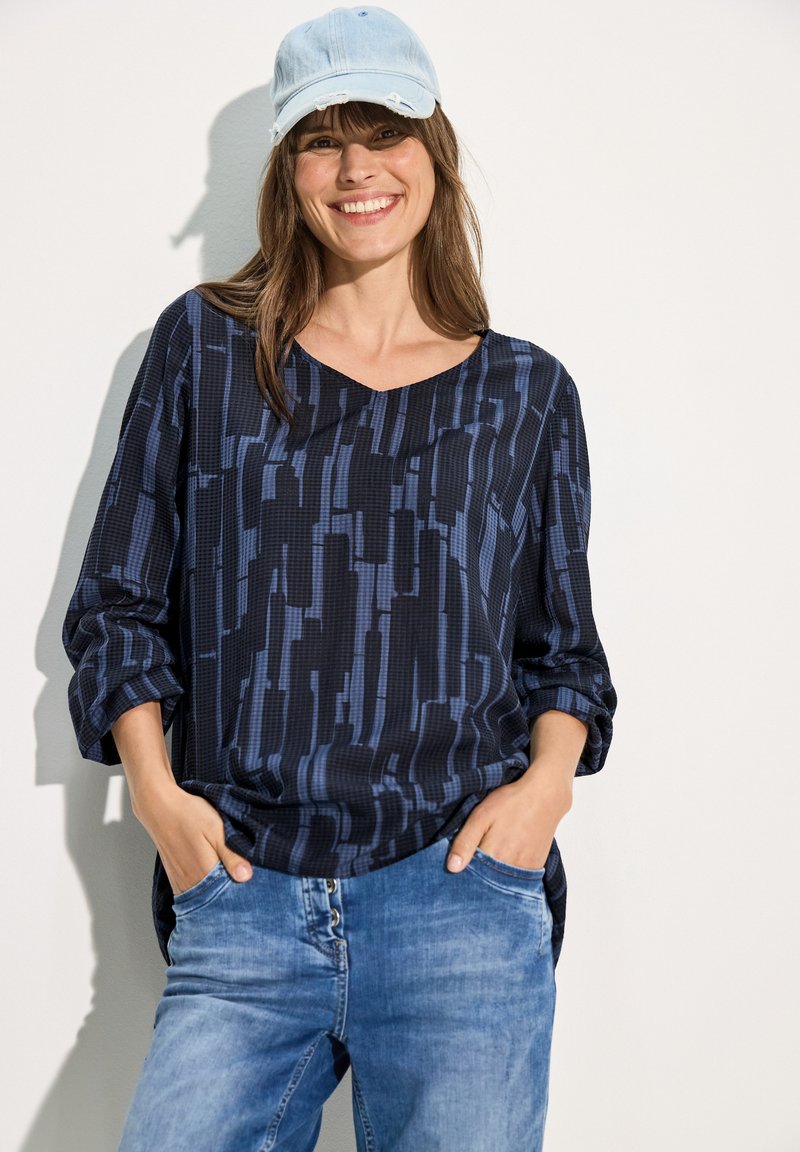 Dunkelblaues gemustertes Oberteil mit V-Ausschnitt und langen Ärmeln, kombiniert mit hellblauen Jeans. Das Shirt weist geometrische Designs in verschiedenen Farbtönen auf.