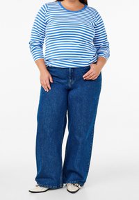 Blå och vitrandig långärmad skjorta tillsammans med vida blå denimjeans och krämfärgade skor mot en enfärgad bakgrund.