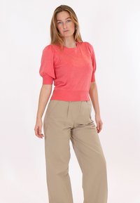 Naine, kes kannab korallikarva volangiga varrukatega tippi ja beežid kõrge vöökohaga laia säärega pükse, seisab ühevärvilise valge tausta ees.