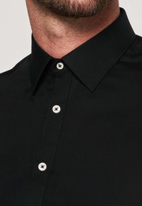 Camicia nera in cotone con colletto classico, caratterizzata da bottoni bianchi e un tessuto liscio e strutturato. Vista ravvicinata del collo e della parte superiore del torso.