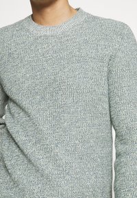 Pull en maille bleu clair et gris avec un col rond, présentant un motif texturé et des poignets côtelés. Coupe décontractée, avec une finition en tissu doux.