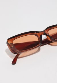 Ochelari de soare dreptunghiulari, translucizi, de culoare maro, cu lentile în nuanță portocalie și brațe elegante. Prezintă un logo subtil al brandului pe rama interioară.