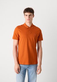 Oransje polo skjorte med krage, to knapper foran og et lite logo-patch, kombinert med lyseblå denim jeans.