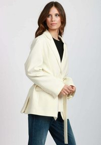 Blazer sartorial di colore giallo chiaro con maniche lunghe, ampi revers e cintura in vita. Presenta una texture liscia e chiusura con un solo bottone. Abbinato a jeans scuri.