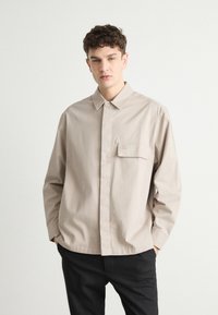 Beige button-up shirt in lightweight fabric, featuring a chest pocket with logo, a fold-down collar, and relaxed fit. Paired with black trousers.