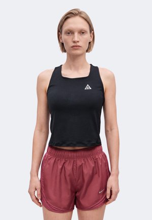 Nainen pukeutuneena mustaan hihattomaan crop-toppiin, jossa on ACG-logo, ja punaisiin urheilushortseihin, seisomassa yksiväristä taustaa vasten.