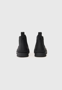 Botas pretas de tornozelo com parte superior sintética lisa, alças de puxar e sola de borracha texturizada. Apresenta um design minimalista sem logótipos visíveis.