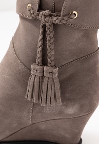 Šedá zamšová kotníková bota s pleteným detailem třásní, zlatými kovovými akcenty a klínovým podpatkem. Hladká textura a elegantní design.